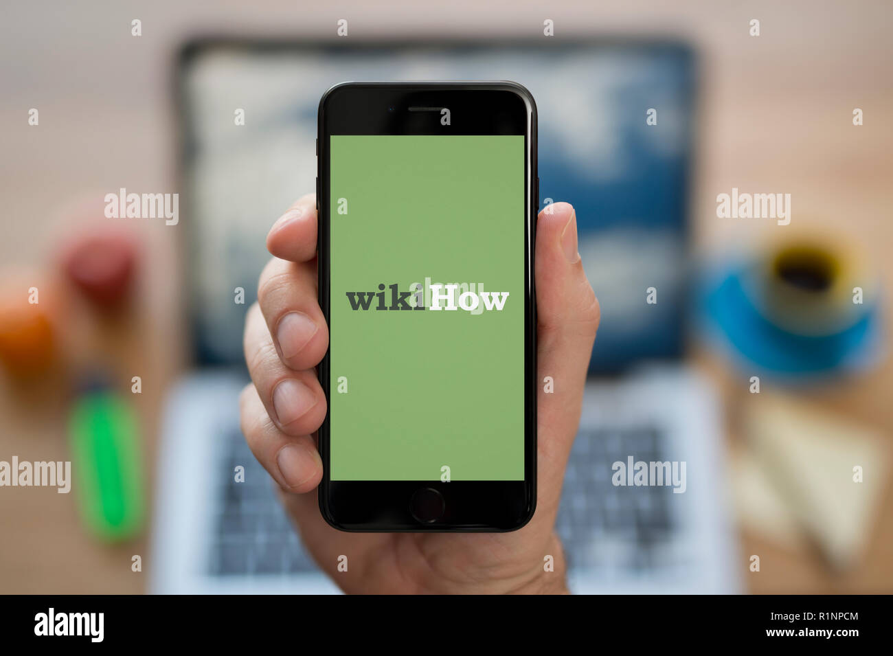 A man looks at his iPhone which displays the WikiHow logo, while sat at his computer desk (Editorial use only). Stock Photo