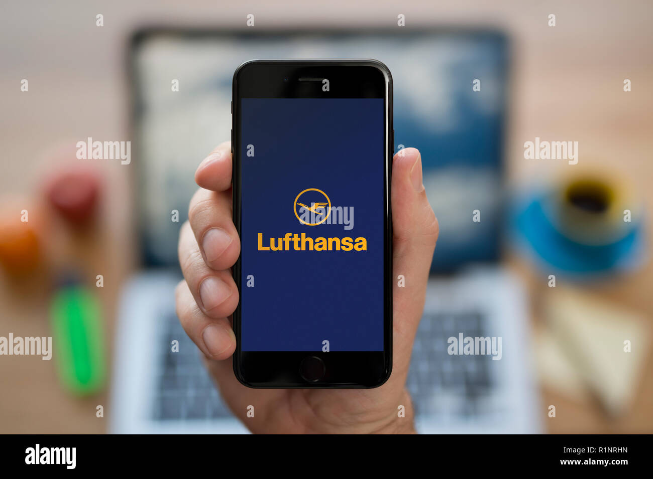 A man looks at his iPhone which displays the Lufthansa logo, while sat at his computer desk (Editorial use only). Stock Photo