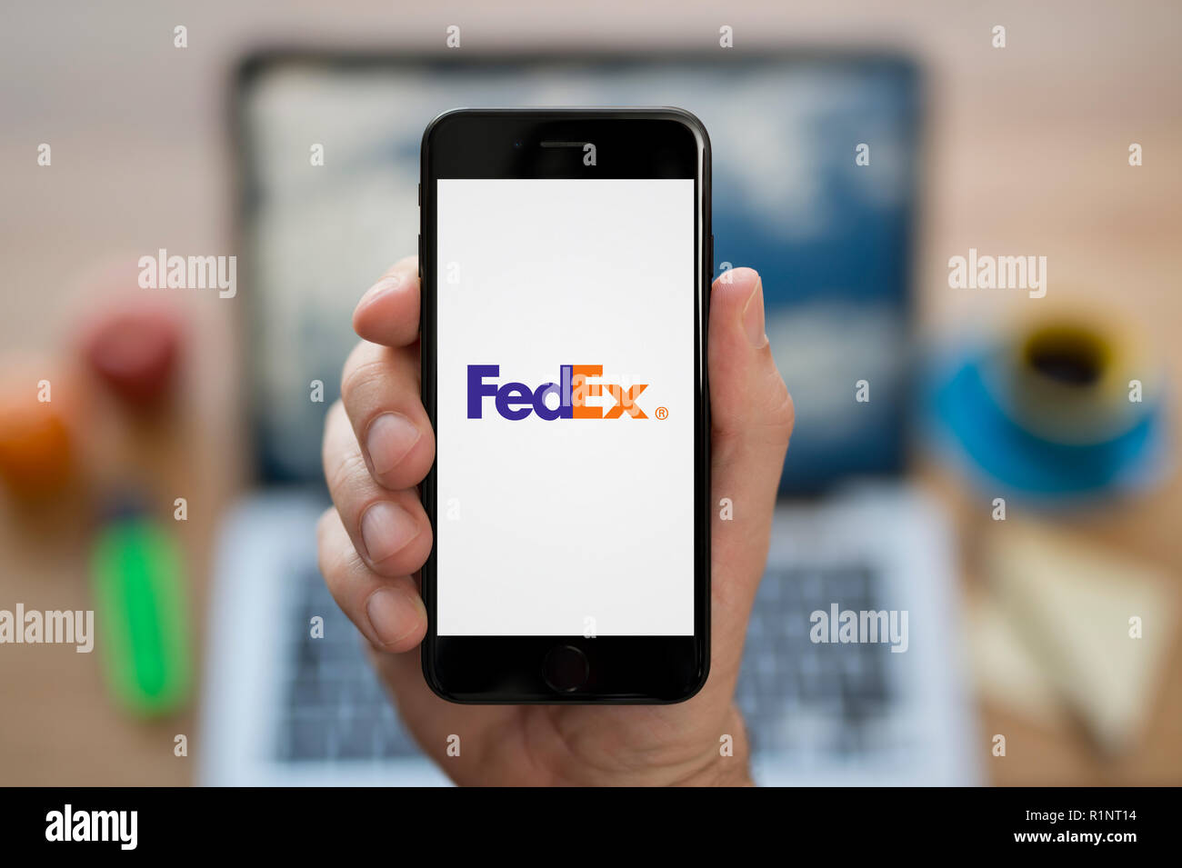 A man looks at his iPhone which displays the Fed Ex logo, while sat at his computer desk (Editorial use only). Stock Photo