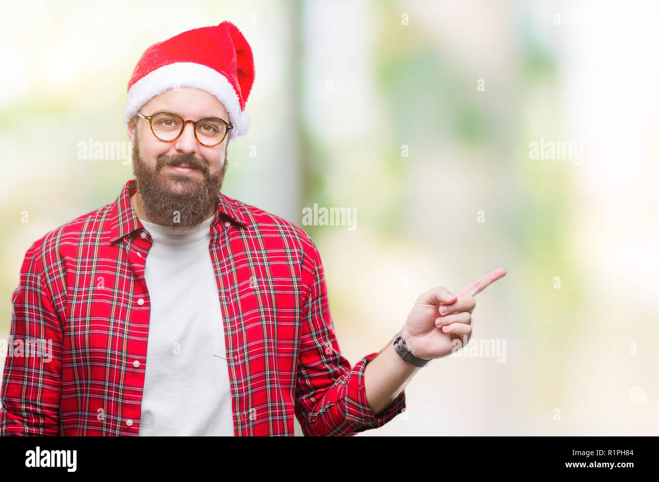 Young caucasian man wearing christmas hat over isolated background with a big smile on face, pointing with hand and finger to the side looking at the  Stock Photo