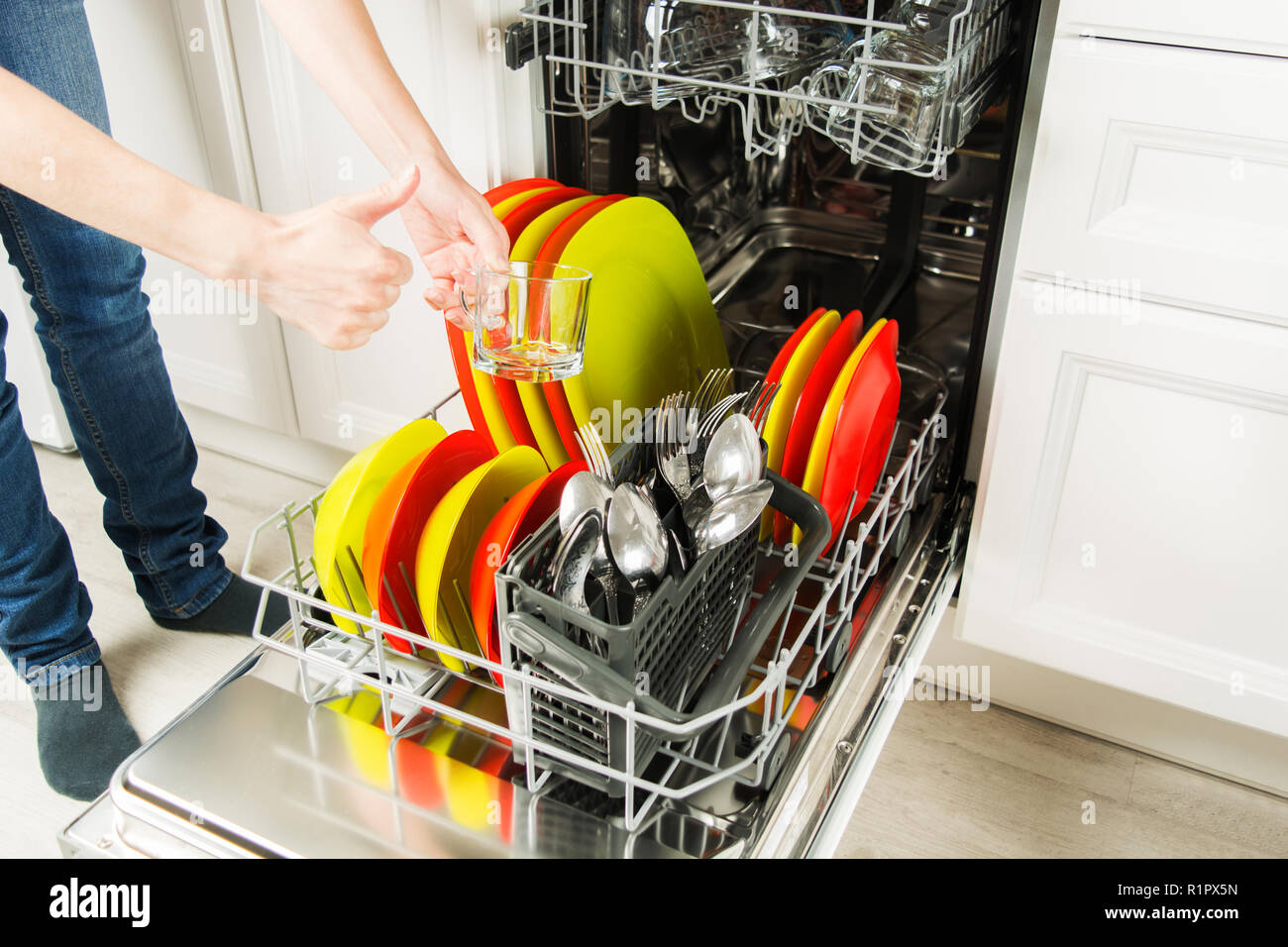 Female legs in jeans with hands showing thumbs up for clean plate after dishwasher, housework concept Stock Photo