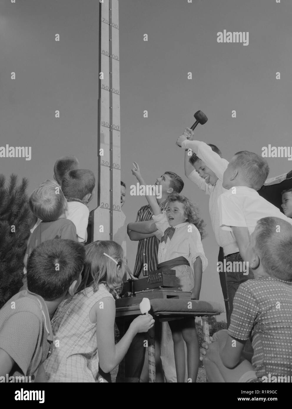 Children gather around a bell ringing carnival game, ca. 1957. Stock Photo