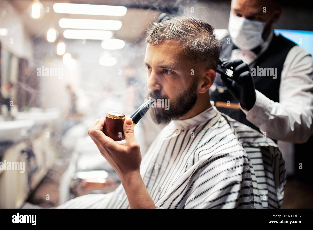 Hipster man client visiting haidresser and hairstylist in barber shop, smoking a pipe. Stock Photo