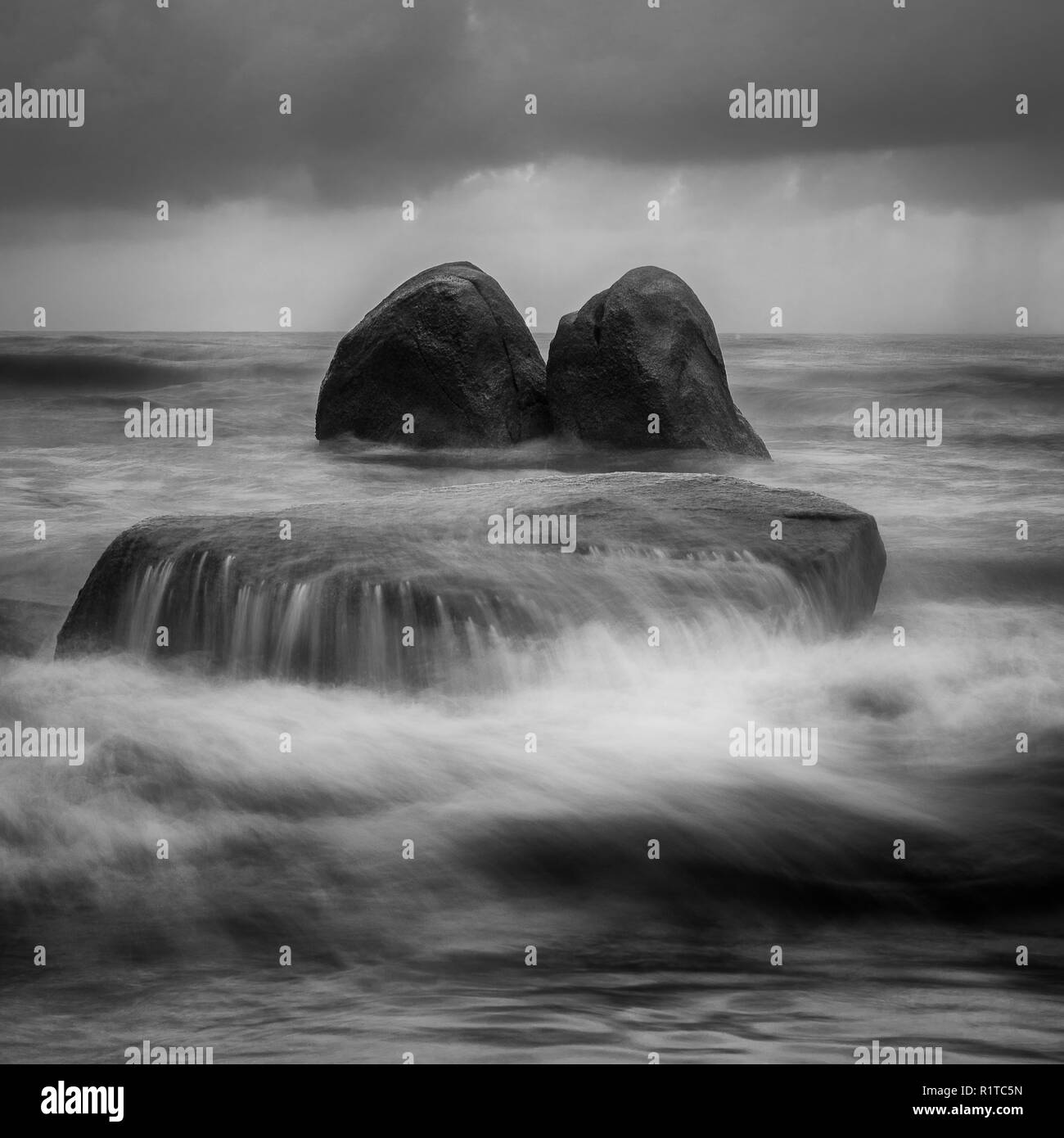 Artistic black and white landscape image of rock being crashed by the waves at the beach with dark cloud before rain. Stock Photo