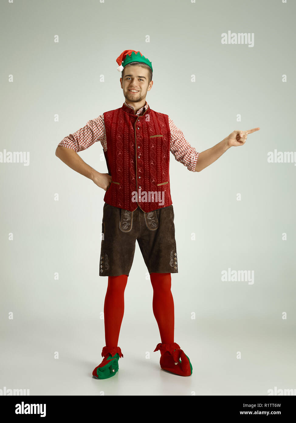 The happy smiling friendly man dressed like a funny gnome or elf pointing to left on an isolated gray studio background. The winter, holiday, christmas concept Stock Photo