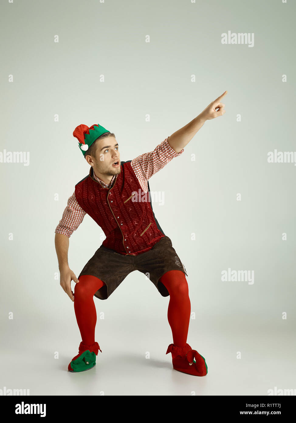 The happy smiling friendly man dressed like a funny gnome or elf pointing to left on an isolated gray studio background. The winter, holiday, christmas concept Stock Photo