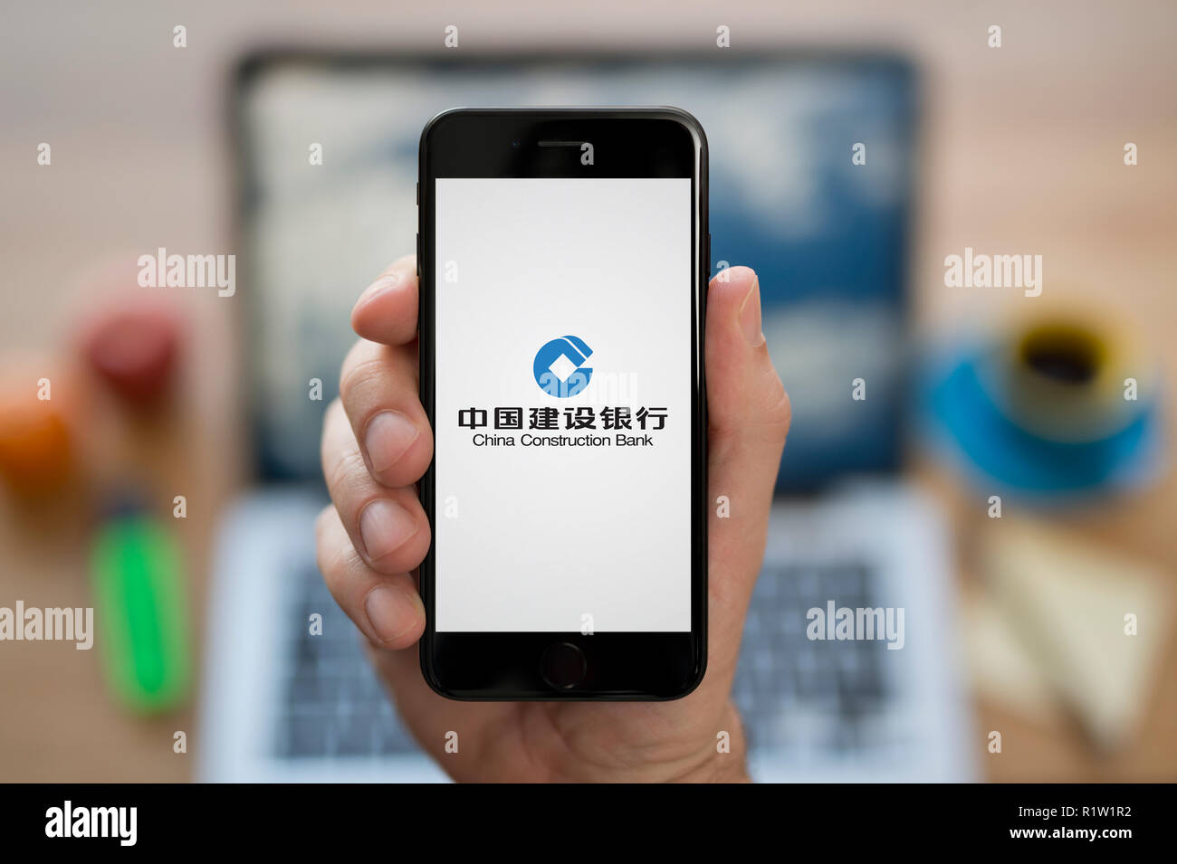 A man looks at his iPhone which displays the China Construction Bank logo, while sat at his computer desk (Editorial use only). Stock Photo