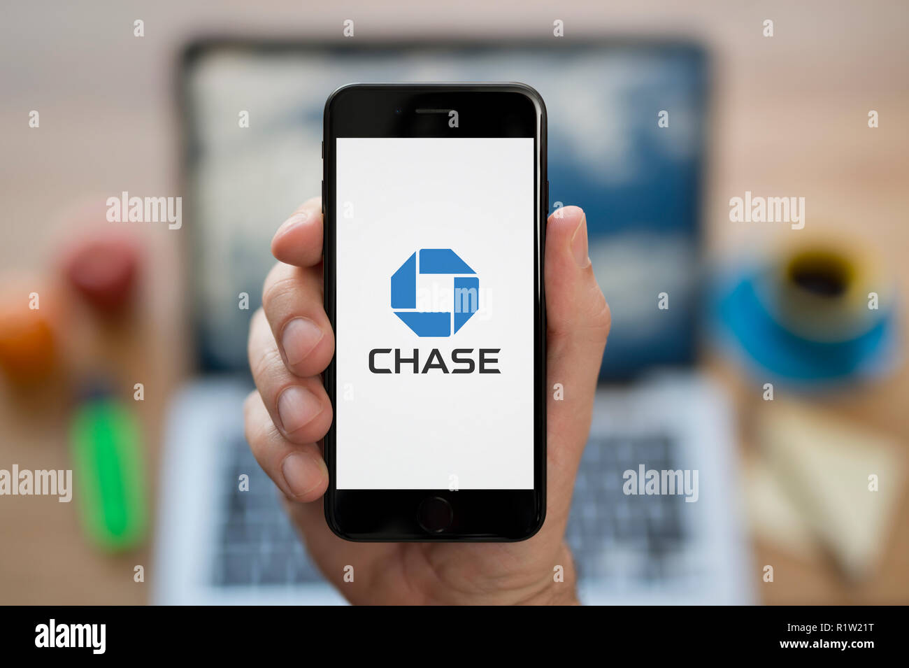 A man looks at his iPhone which displays the Chase bank logo, while sat at his computer desk (Editorial use only). Stock Photo