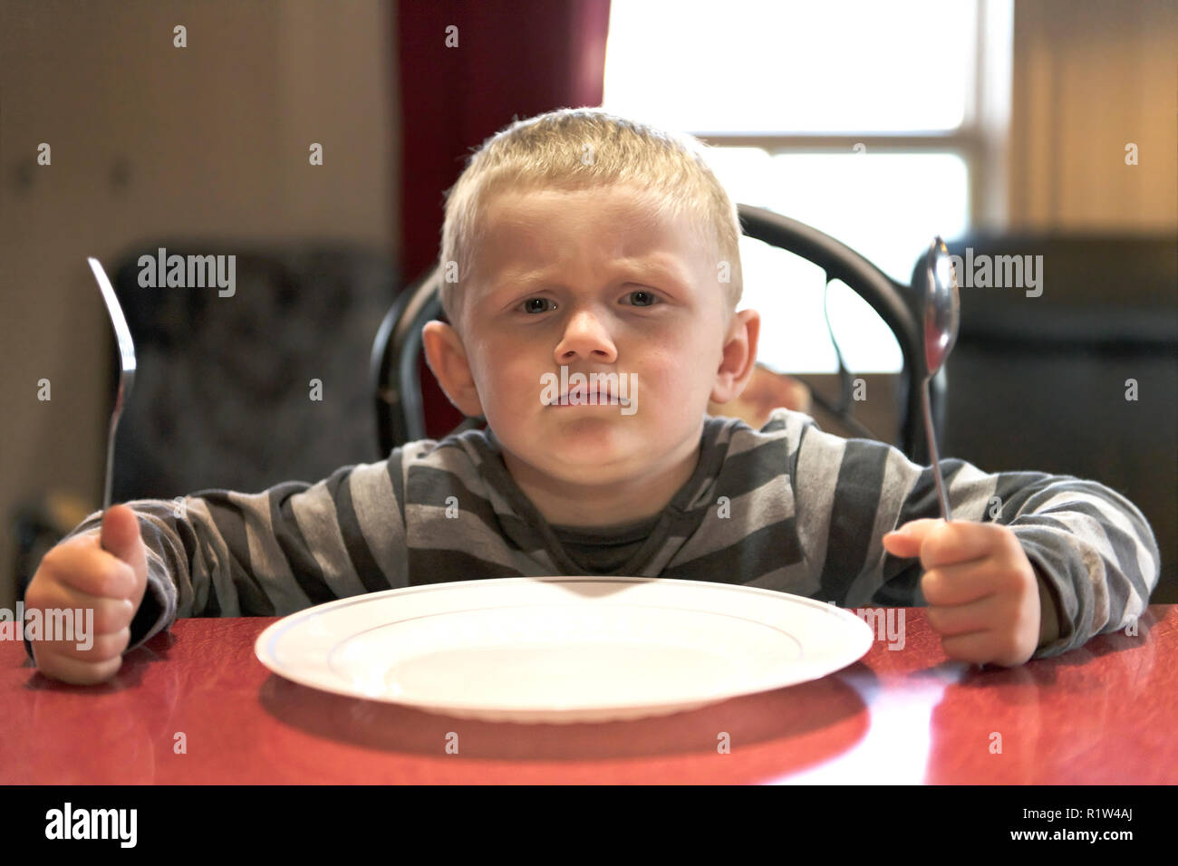 Upset little boy waiting for dinner while holding a fork and a spoon Stock Photo