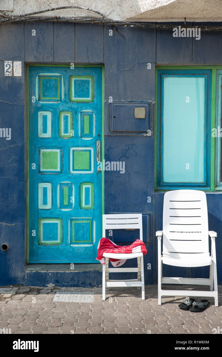 Two empty chairs on the seafront against a blue wall and blue and green door, the chairs are empty, with a beach towel. People have gone swimming Stock Photo