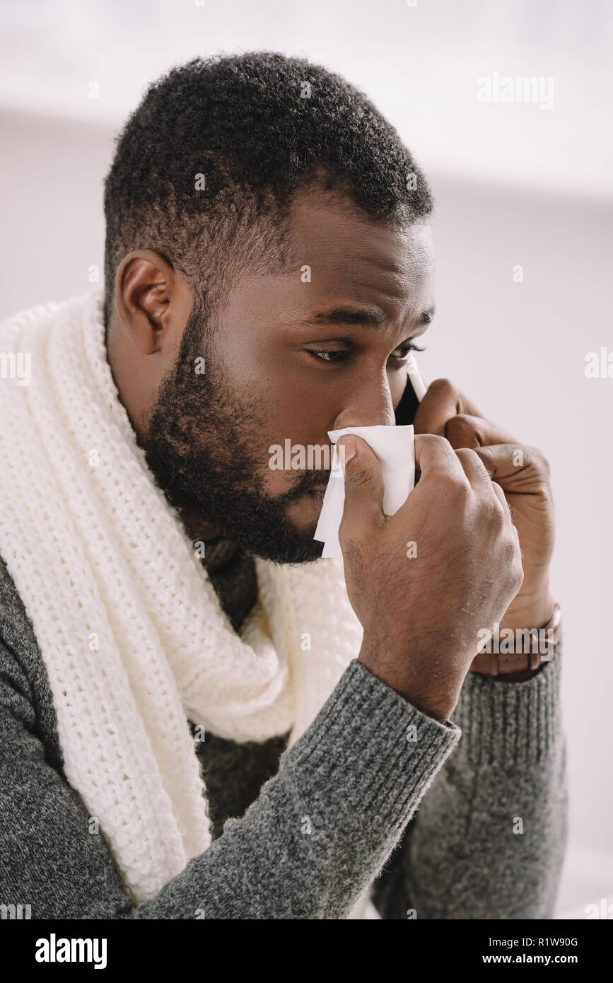 cold african american man in warm scarf with runny nose holding napkin while talking on smartphone Stock Photo