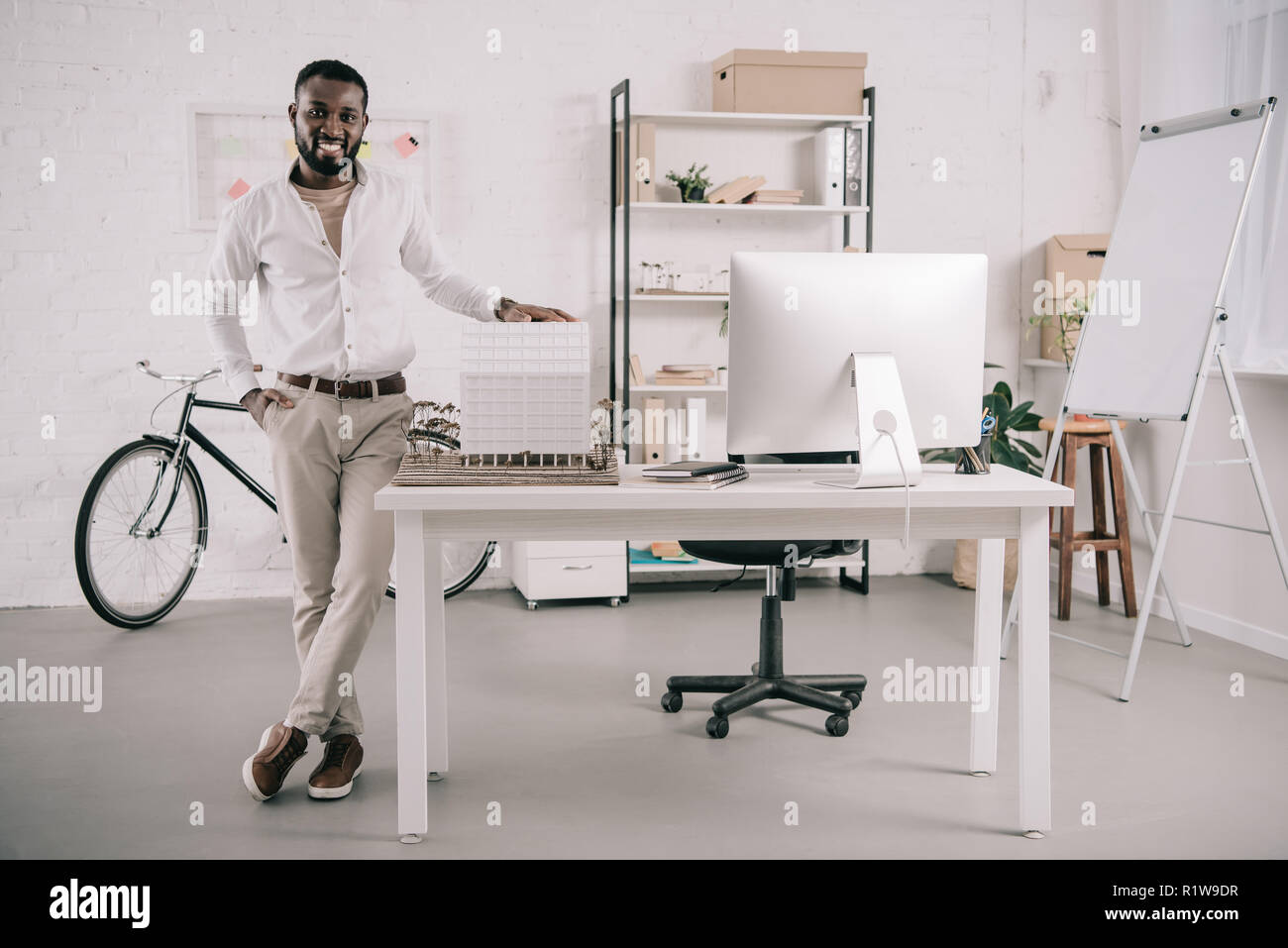 cheerful handsome african american architect touching architecture model and looking at camera in office Stock Photo