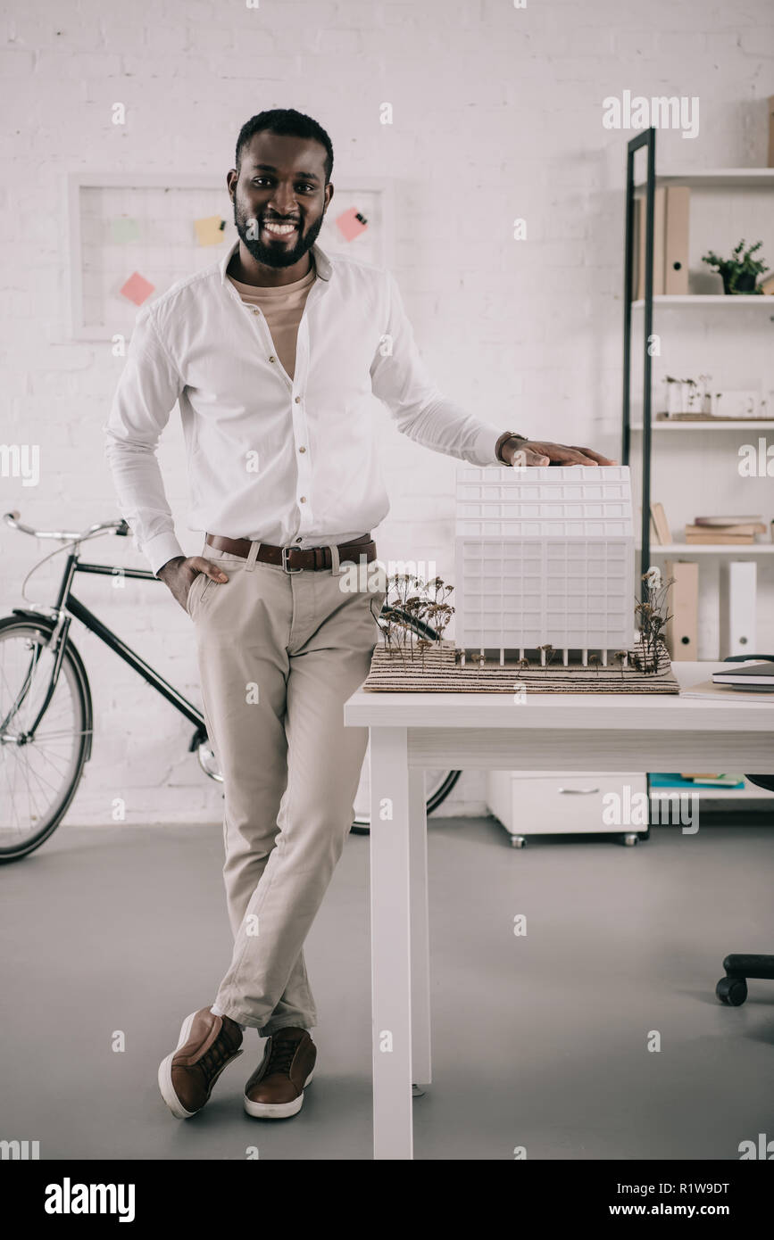 smiling handsome african american architect touching architecture model and looking at camera in office Stock Photo
