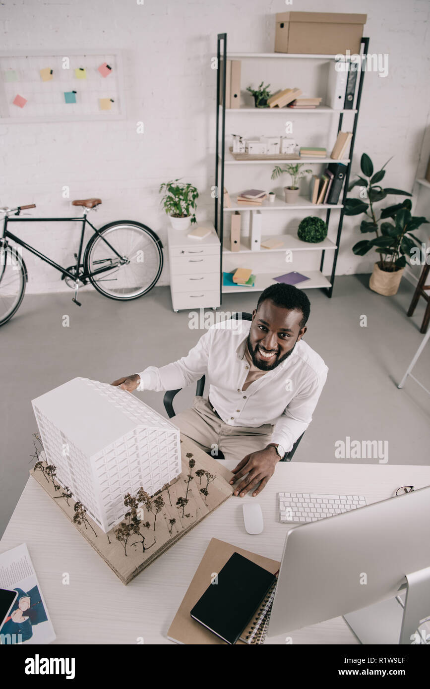 high angle view of smiling handsome african american architect sitting near architecture model and looking at camera in office Stock Photo
