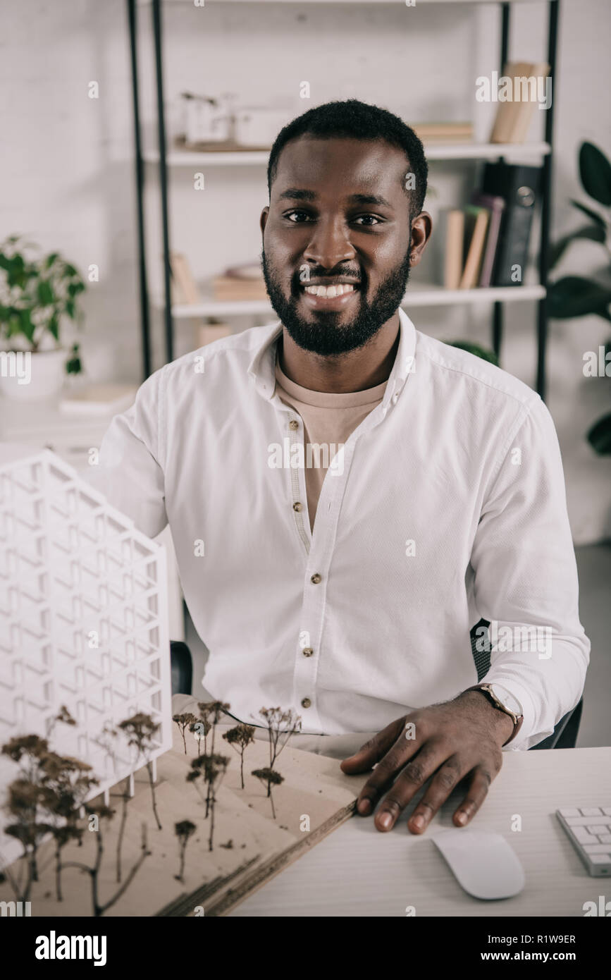 smiling african american architect sitting near architecture model on table in office and looking at camera Stock Photo