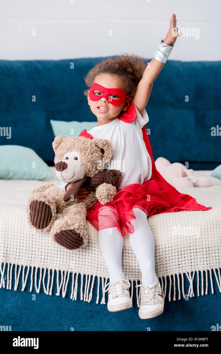 Adorable little african american child in superhero costume sitting on bed with teddy bear Stock Photo