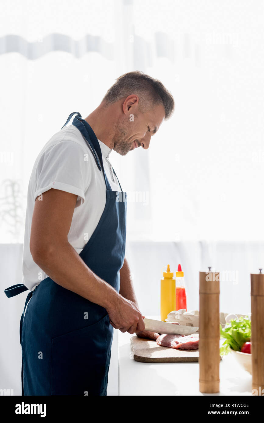 Premium Photo  Man cuts of fresh piece of meat on a wooden cutting board  in the home kitchen. a man in a striped apron with a big knife in his hands