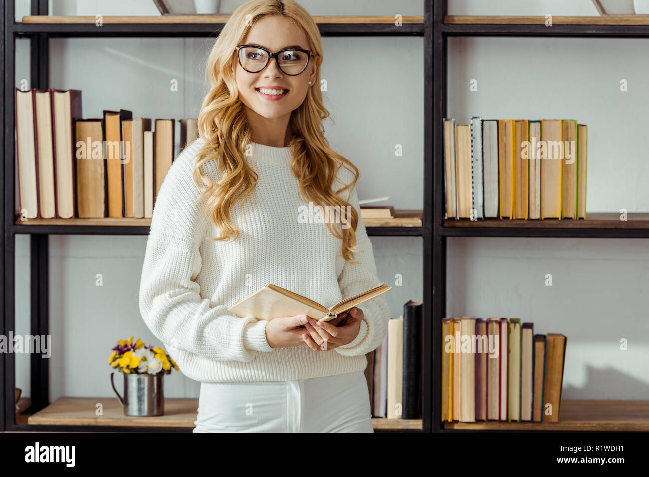 close up of cheerful woman in glasses with book standing near rack Stock Photo