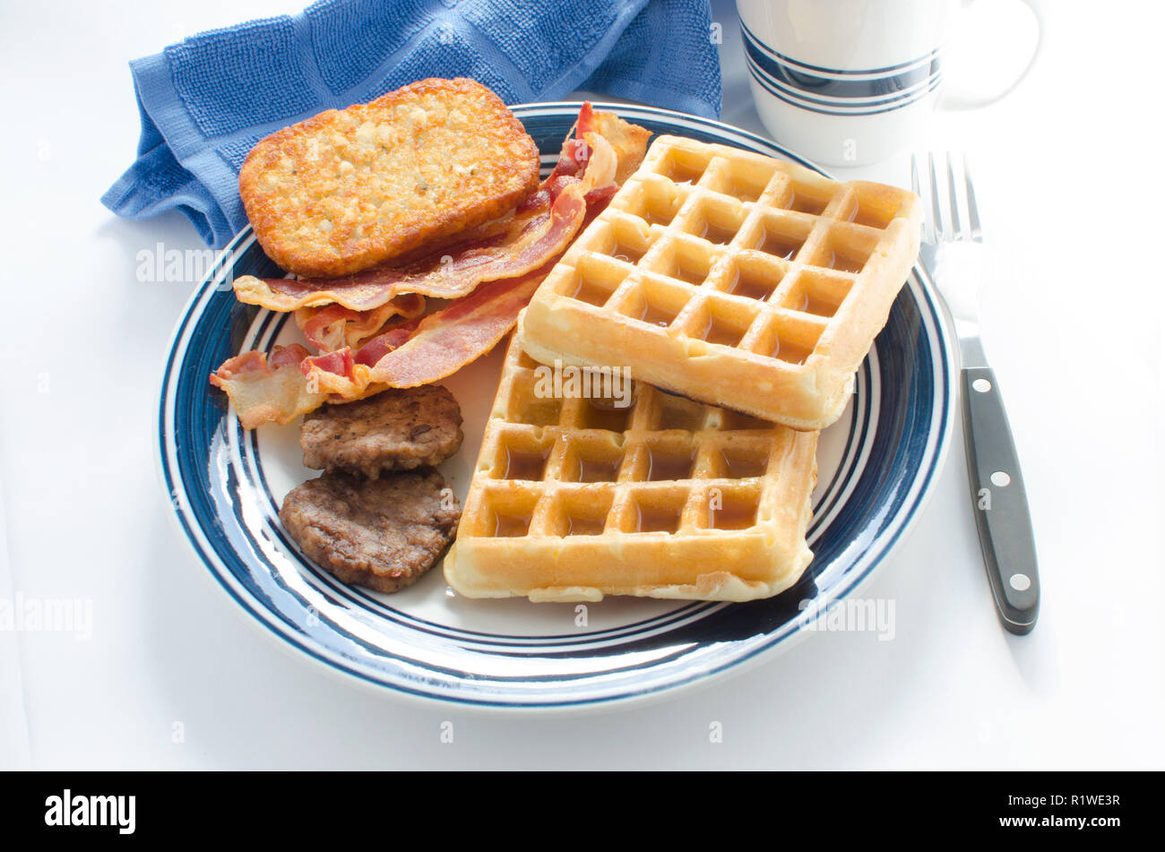 Plate of waffles with maple syrup, bacon strips, sausage patties and a hash brown potato Stock Photo