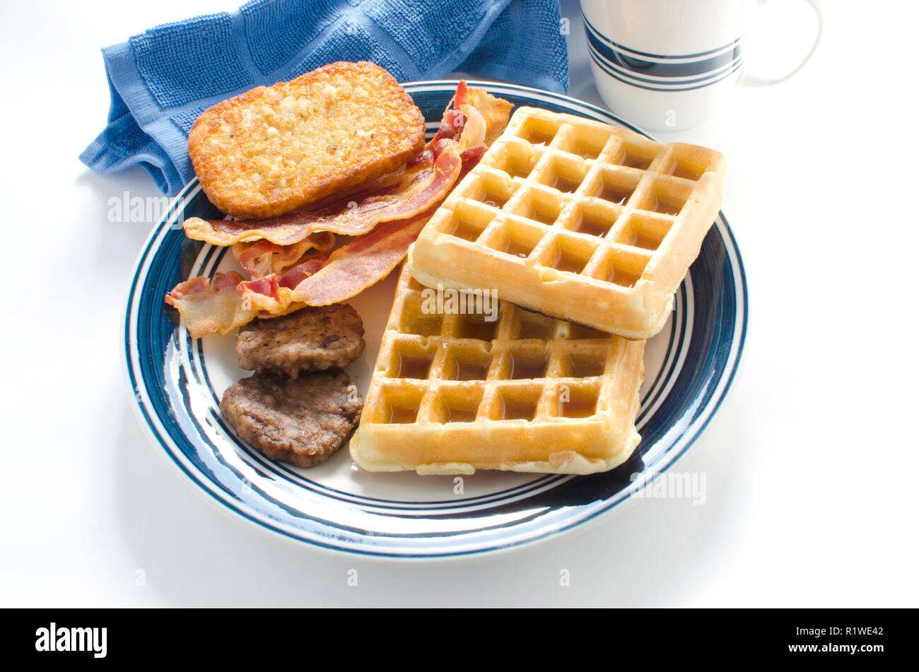 Plate of waffles with maple syrup, sausage patties, bacon strips and a hash brown potato Stock Photo