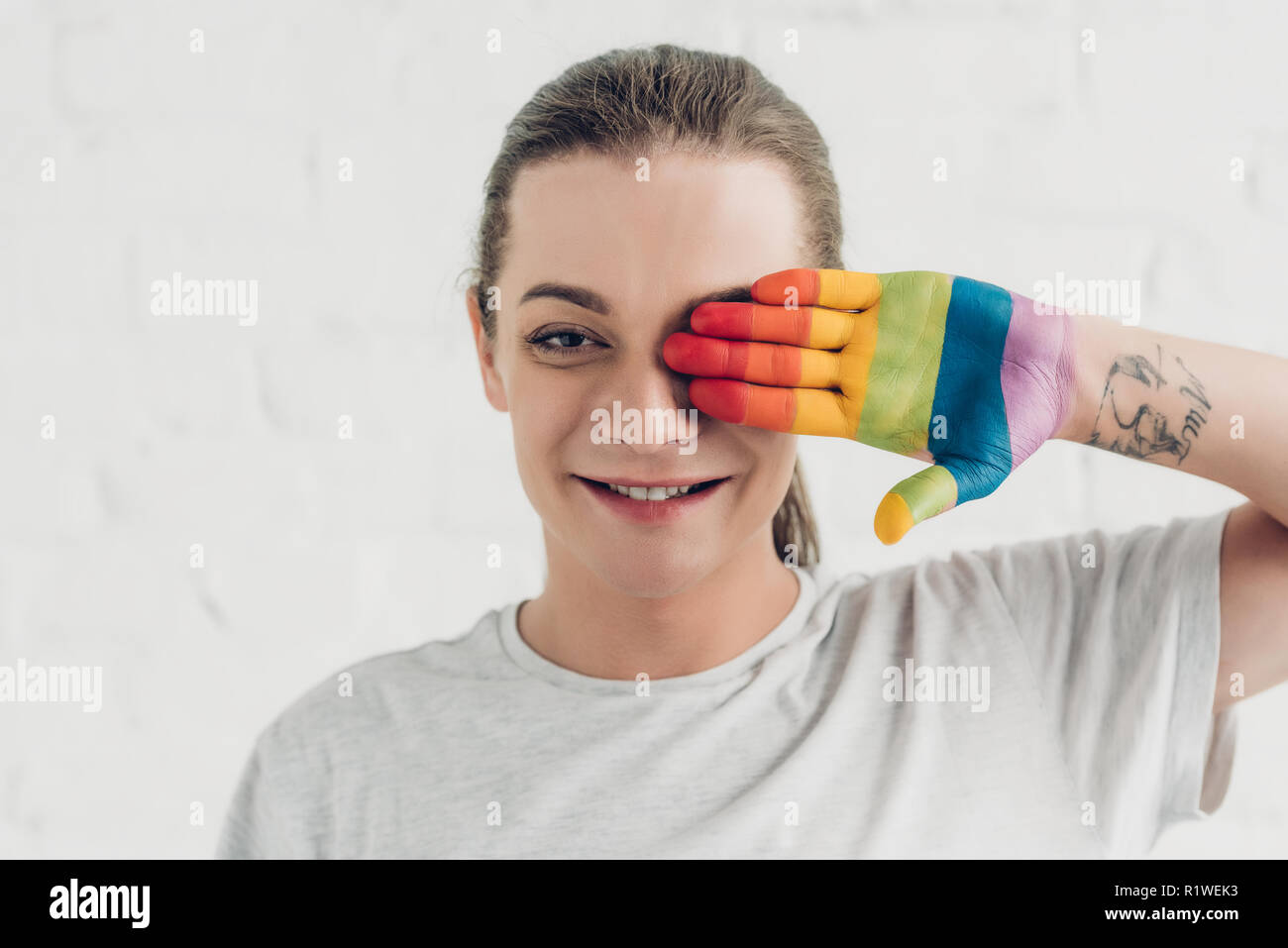 young transgender man covering eye with hand painted in colors of pride flag in front of white brick wall Stock Photo