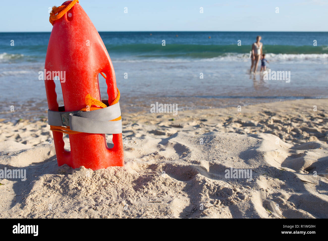 Orange rescue buoy planted on sand beach. Sea weaves as background. Mother with little boy at the bottom Stock Photo