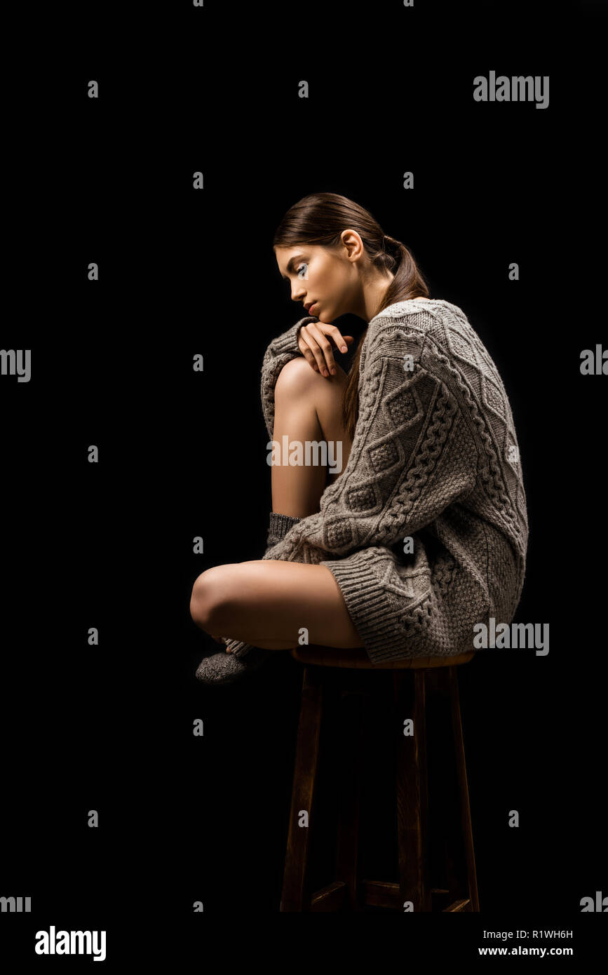 side view of beautiful thoughtful woman in woolen grey sweater sitting on bar stool isolated on black Stock Photo