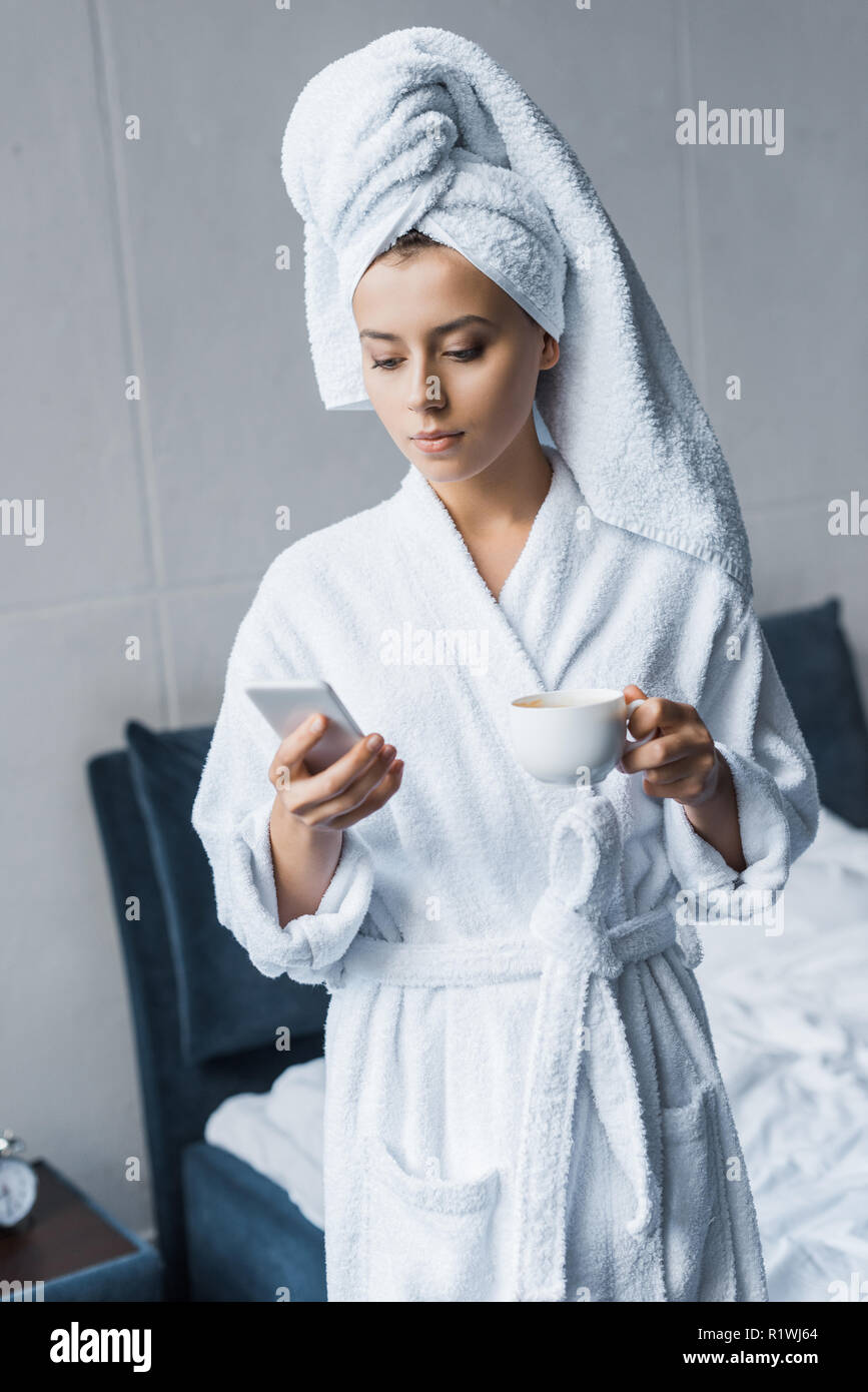 young woman in white bathrobe and towel holding cup of coffee and using  smartphone in bedroom Stock Photo - Alamy