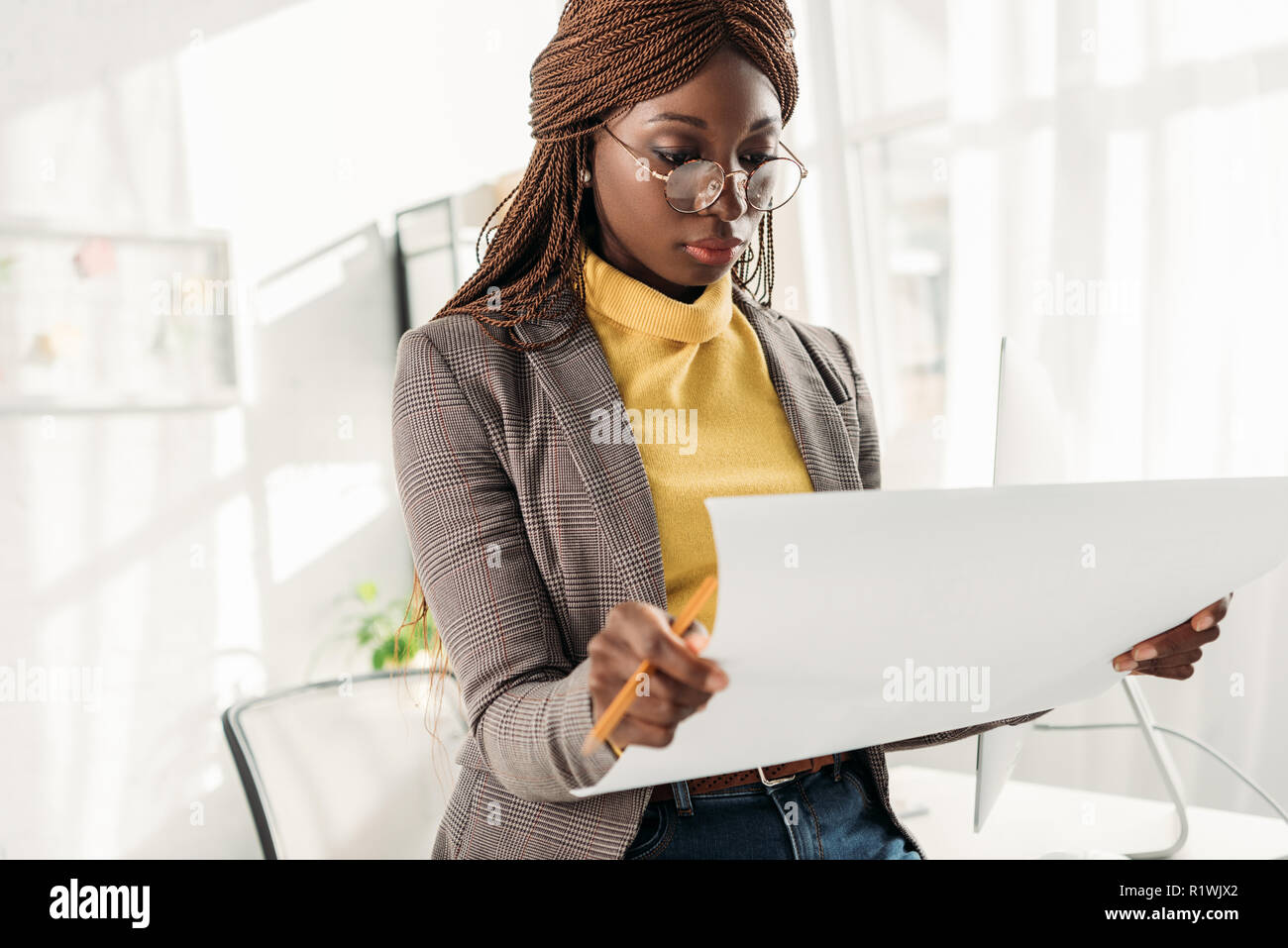 focused african american female architect in glasses holding blueprint and pencil in office Stock Photo