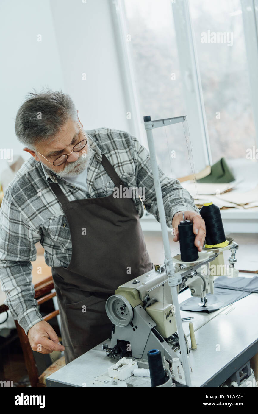 focused middle aged tailor in apron setting strings on sewing machine