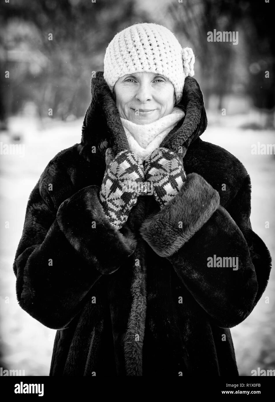 A winter portrait of a smiling senior adult woman wearing a wool cap, a scarf and colored gloves, with a snow background Stock Photo