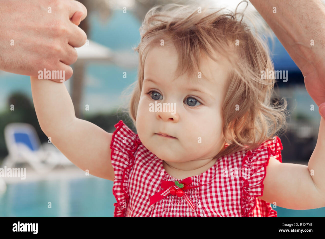 Adorable baby girl holding father's hands while learning to walk outdoors. Swimming pool background. Baby looking up Stock Photo