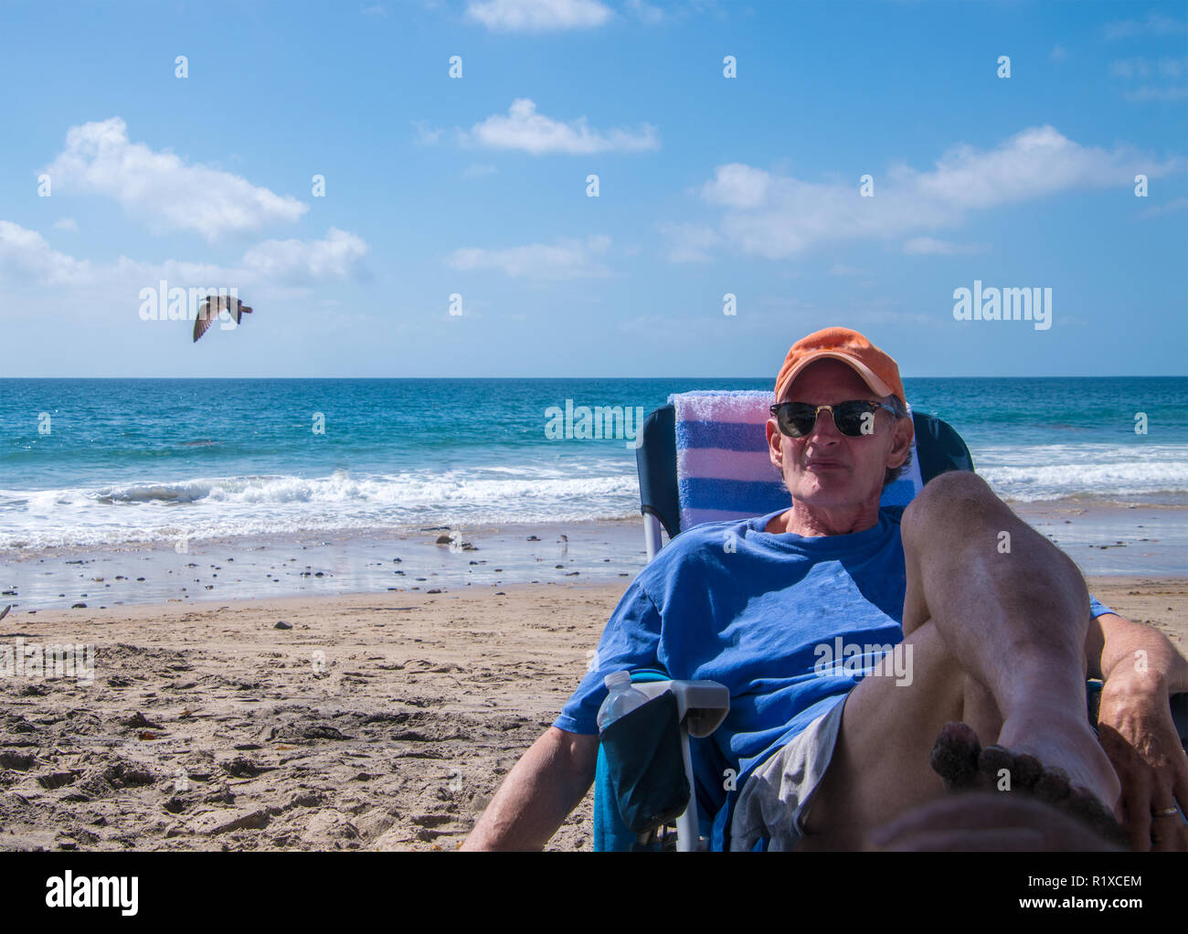 Handsome older baby boomer man wearing an orange cap sitting on beach chair by the ocean with a serious look on his face Stock Photo