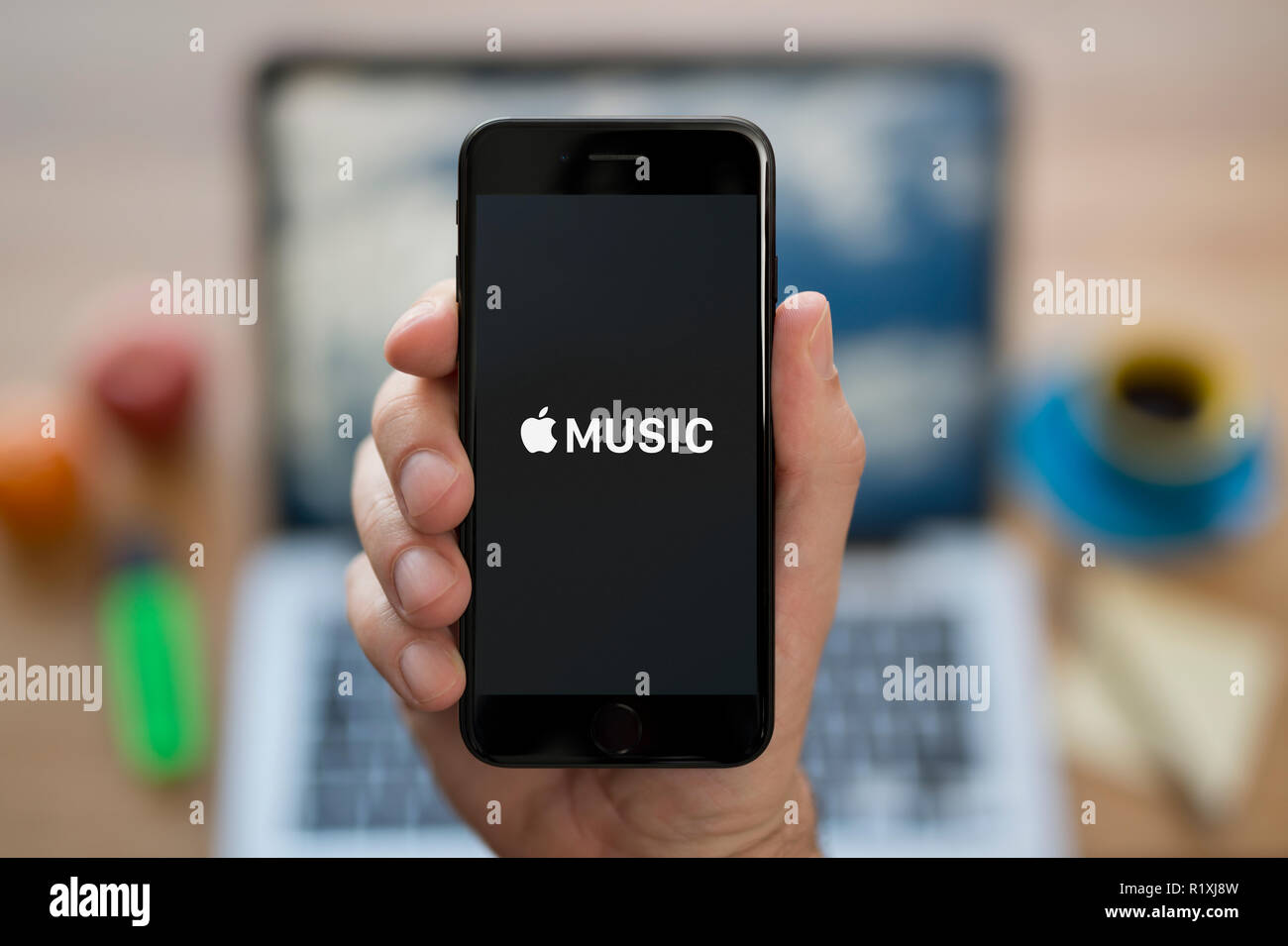 A man looks at his iPhone which displays the Apple Music logo, while sat at his computer desk (Editorial use only). Stock Photo