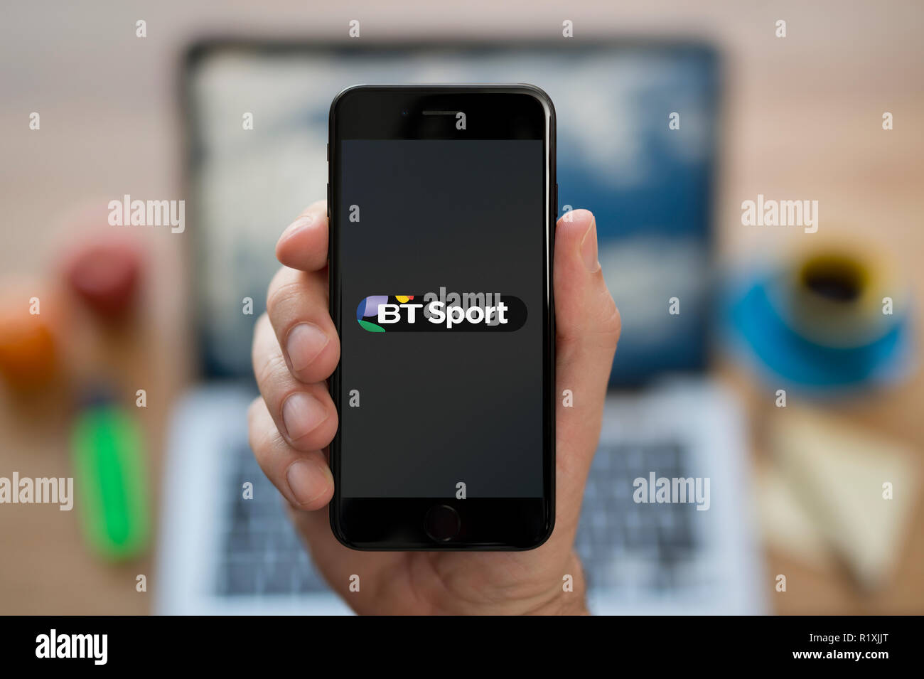 A man looks at his iPhone which displays the BT Sport logo, while sat at his computer desk (Editorial use only). Stock Photo