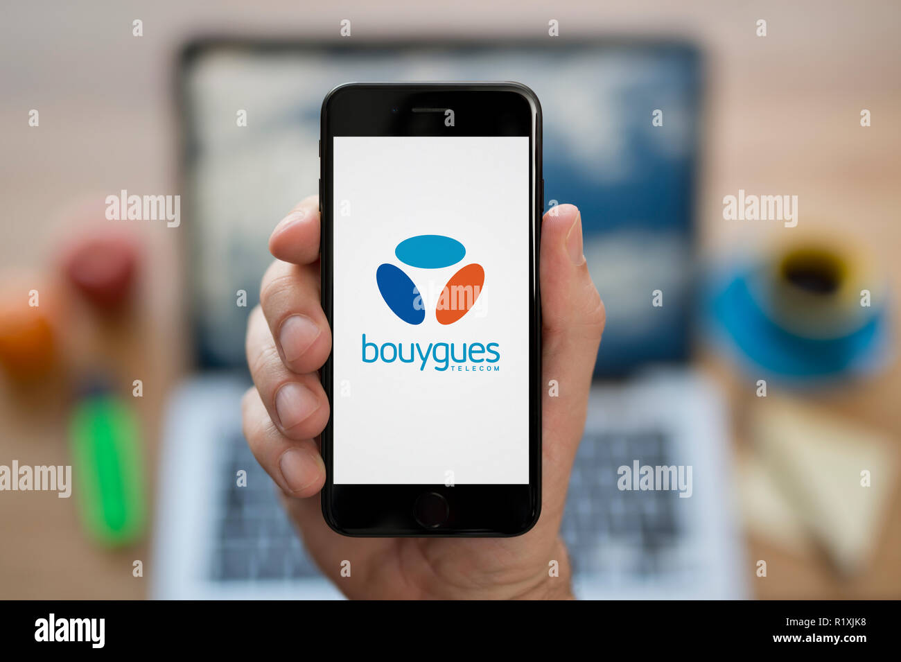 A man looks at his iPhone which displays the Bouygues logo, while sat at his computer desk (Editorial use only). Stock Photo