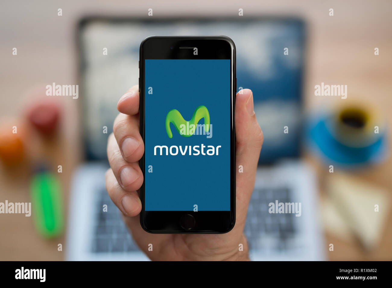 A man looks at his iPhone which displays the Movistar logo, while sat at his computer desk (Editorial use only). Stock Photo