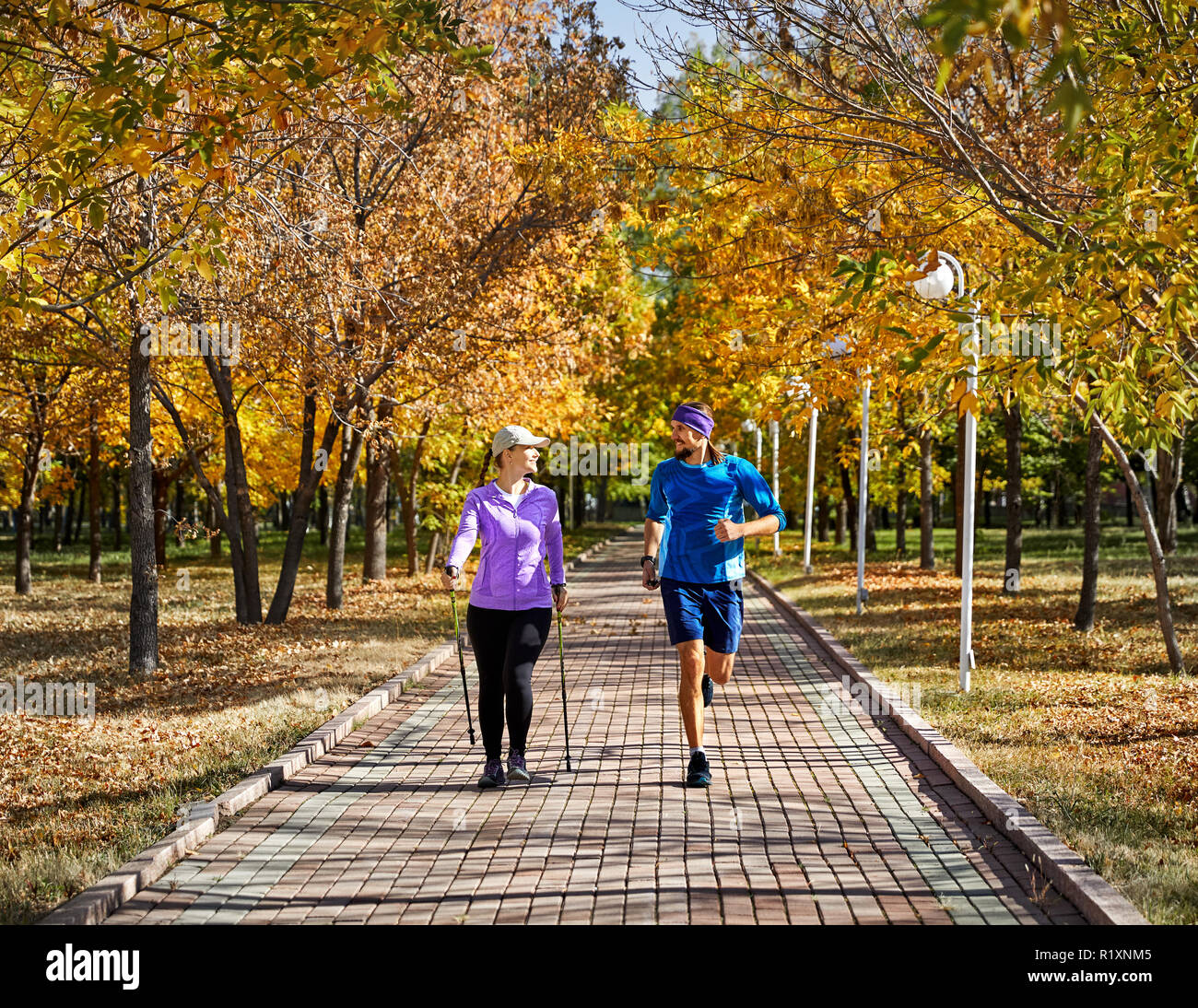 Young couple training in autumn park. Man is jogging and woman doing Nordic walking with poles. Healthy life concept. Stock Photo