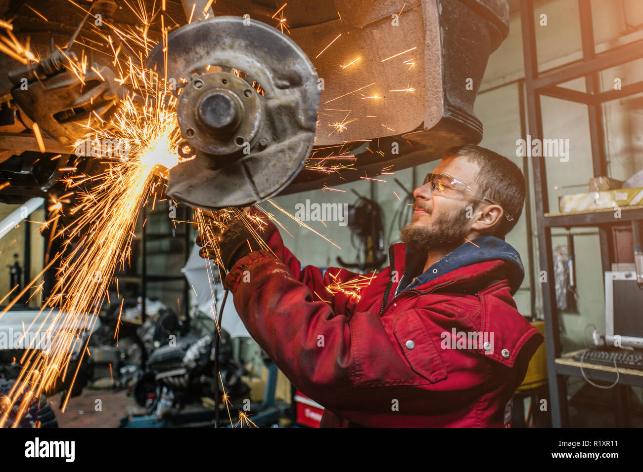 Young caucasian man in red work clothing repairing car with angle cutter. Many sparks on background. Stock Photo