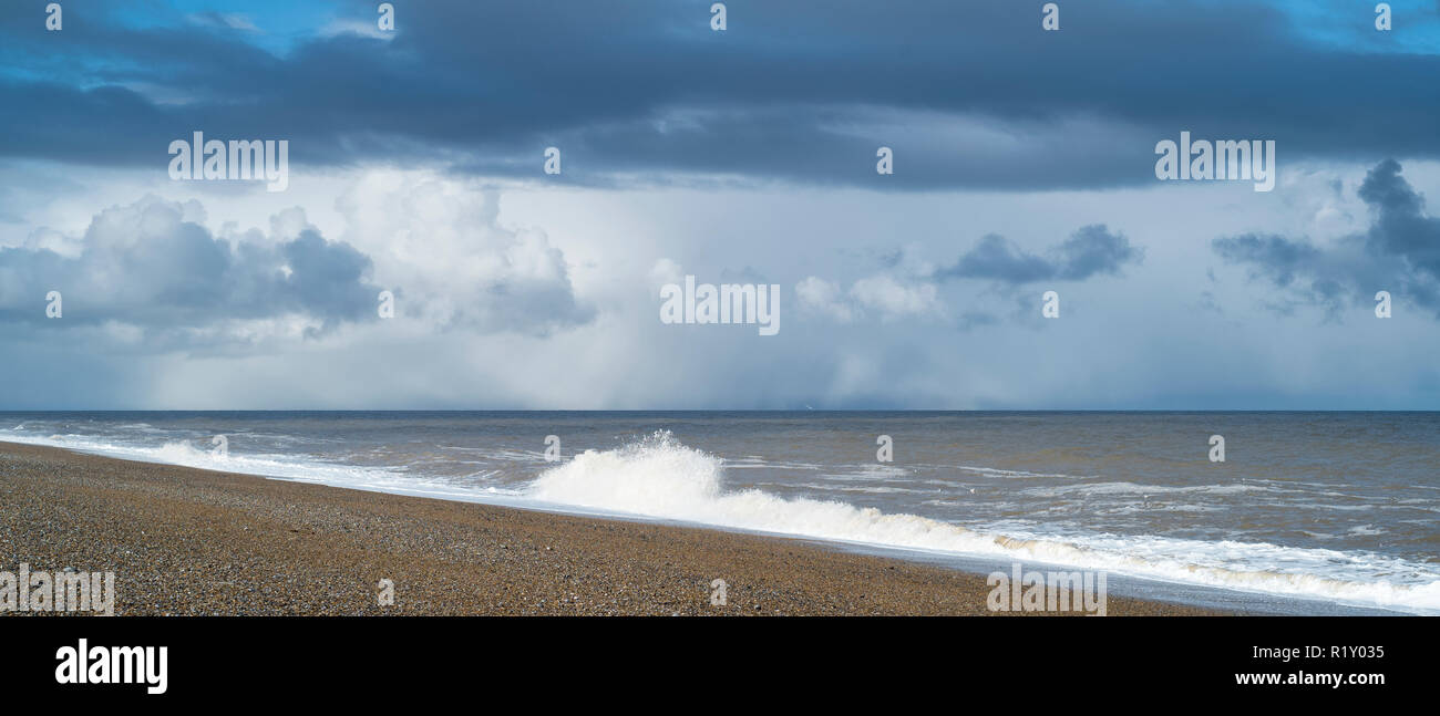 Shoreline, shingle pebble beach and breaking waves under blue sky and puffy dark clouds in wintertime at Cley Next The Sea, Norfolk, UK Stock Photo