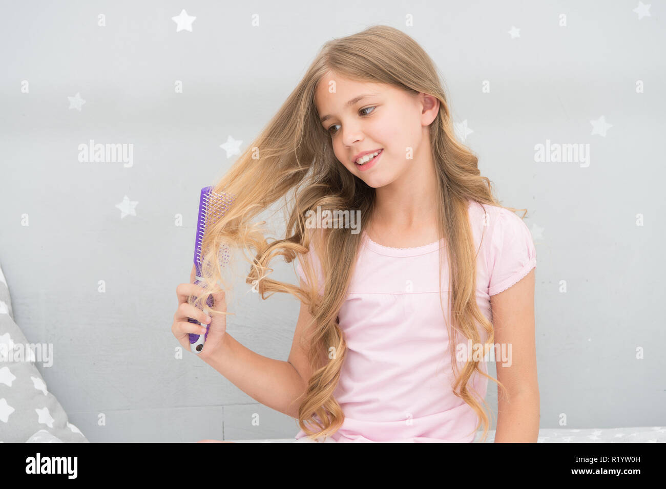 Child curly hairstyle hold hairbrush or comb. Apply oil before combing hair.  Healthy hair. Conditioner or mask organic oil comb hair. Beauty salon tips.  Girl long curly hair grey interior background Stock
