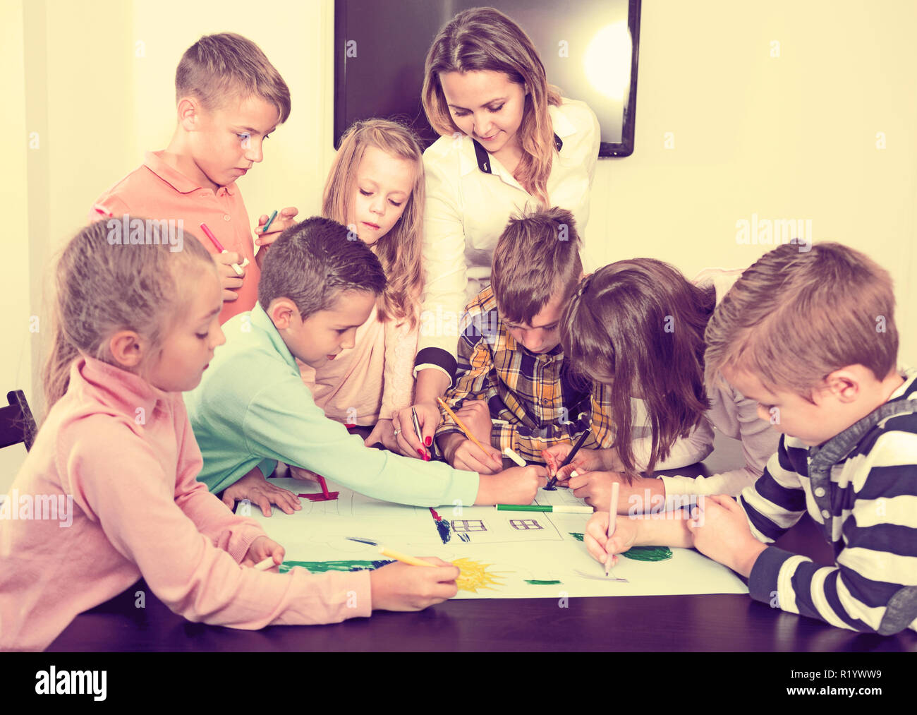 Team of elementary age children and female teacher drawing on one sheet ...