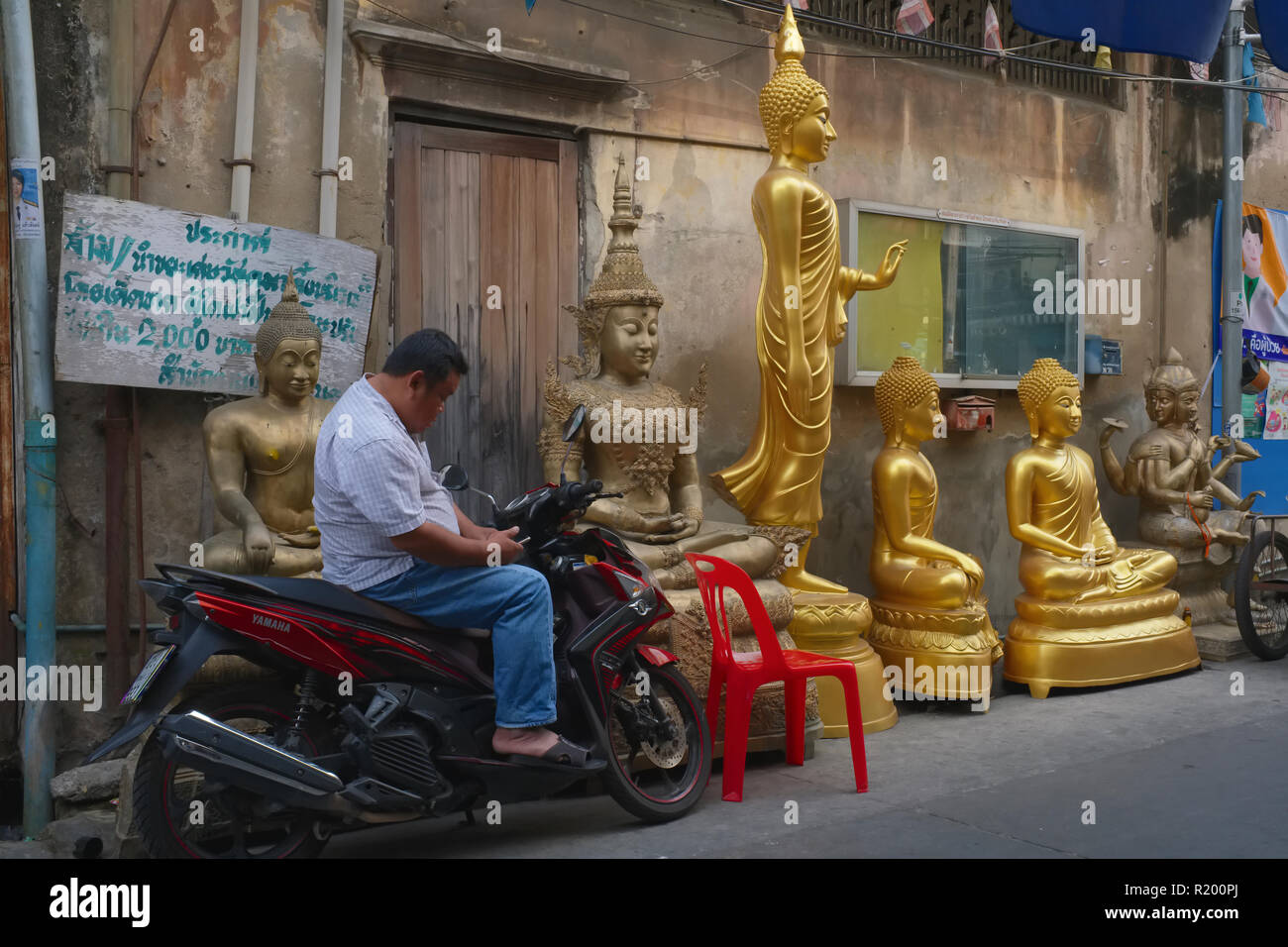 Next to Buddha statues placed outside a Buddha factory in Bamrung Muang Road, Bangkok, Thailand, a man entertains himself with his mobile Stock Photo