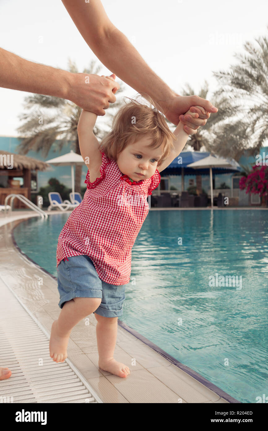 Funny little baby girl near swimming pool. Father holds infant by hands on pool background. Summer vacation with children outdoor Stock Photo