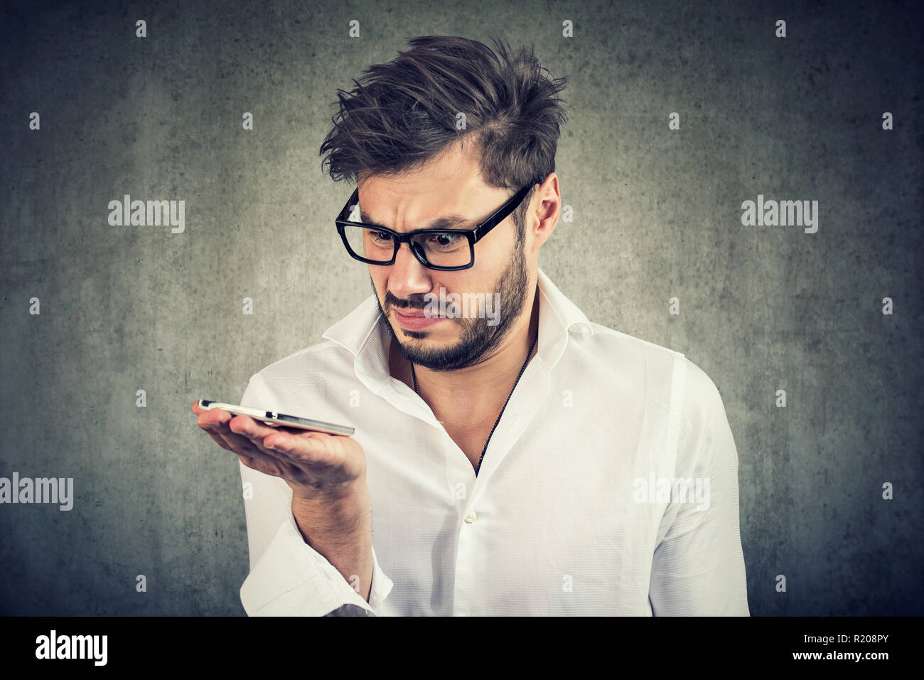 Young bearded man in shirt holding smartphone being upset with voice command function Stock Photo