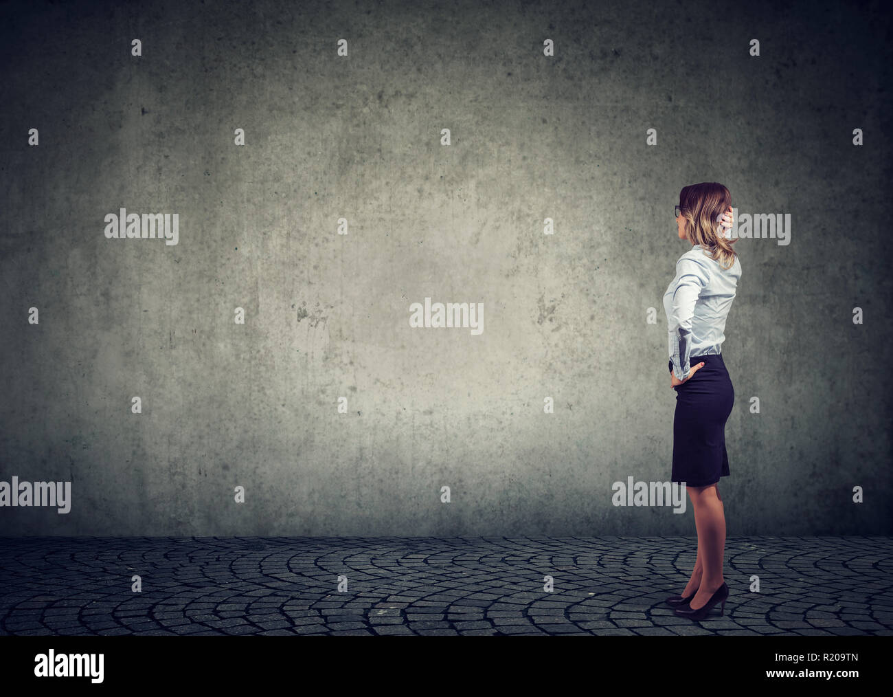 Rear view of business woman standing by the wall with hand on head wondering what to do next being challenged. Full length of businesswoman facing the Stock Photo