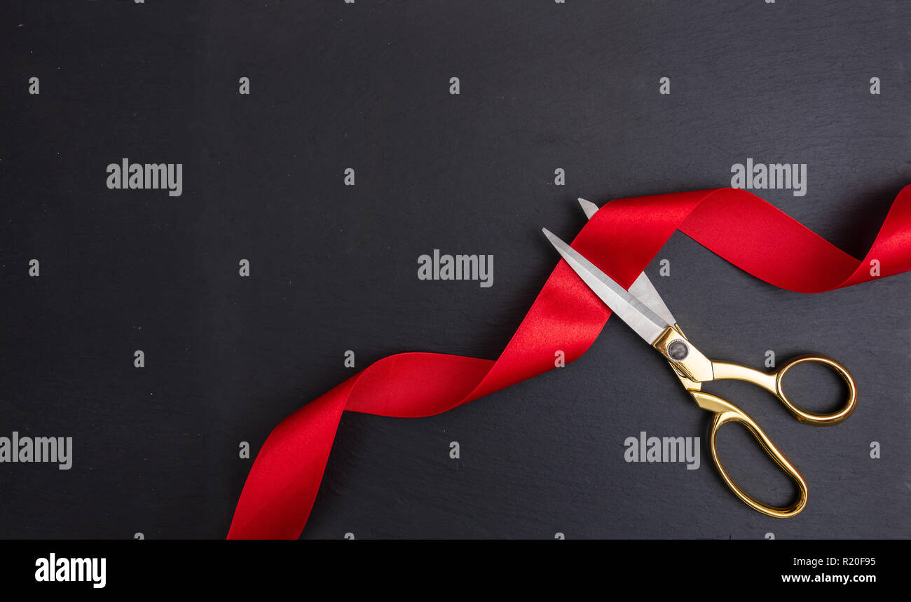 Grand opening. Top view of gold scissors cutting red silk ribbon against black background, copy space Stock Photo