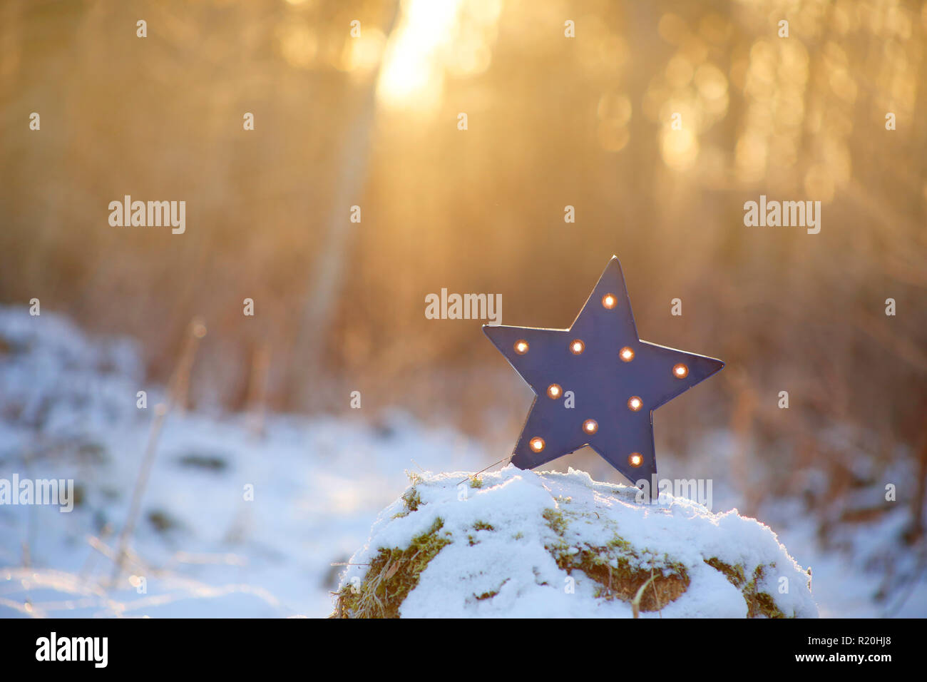 Focus on decorative modern blue star shape lamp with lots of burning small light bulbs on, outdoors on snowy ground, with nice back light from sun set Stock Photo