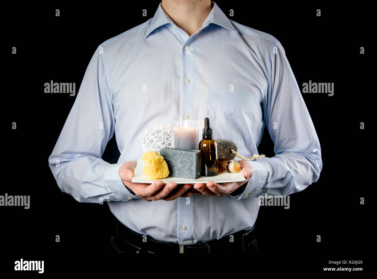 Manly businessman holding white ceramic tray with group of different spa essentials, beard oil, gray soap, natural sea sponge, candle burning, oil. Stock Photo