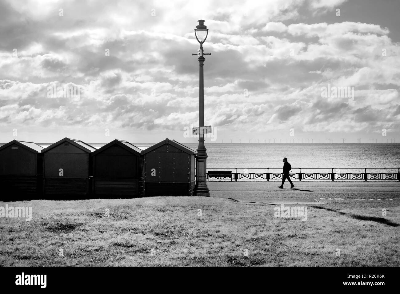 A line of four beach huts and a street lamp silhoutted black by the sun shining directly at the camera through a dramatic blue and white sky, the sea  Stock Photo